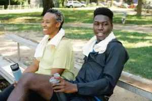 Two adults resting on a park bench with towels and water bottles after exercise.